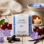 Packshot d'un paquet de quatre carrés fondants Drake à la senteur Alizés devant un brule parfum, des cerises, des fleurs et de la vanille
