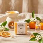Packshot d'un paquet de quatre carrés fondants Drake à la senteur Fleur d'Oranger devant un brule parfum et une orange